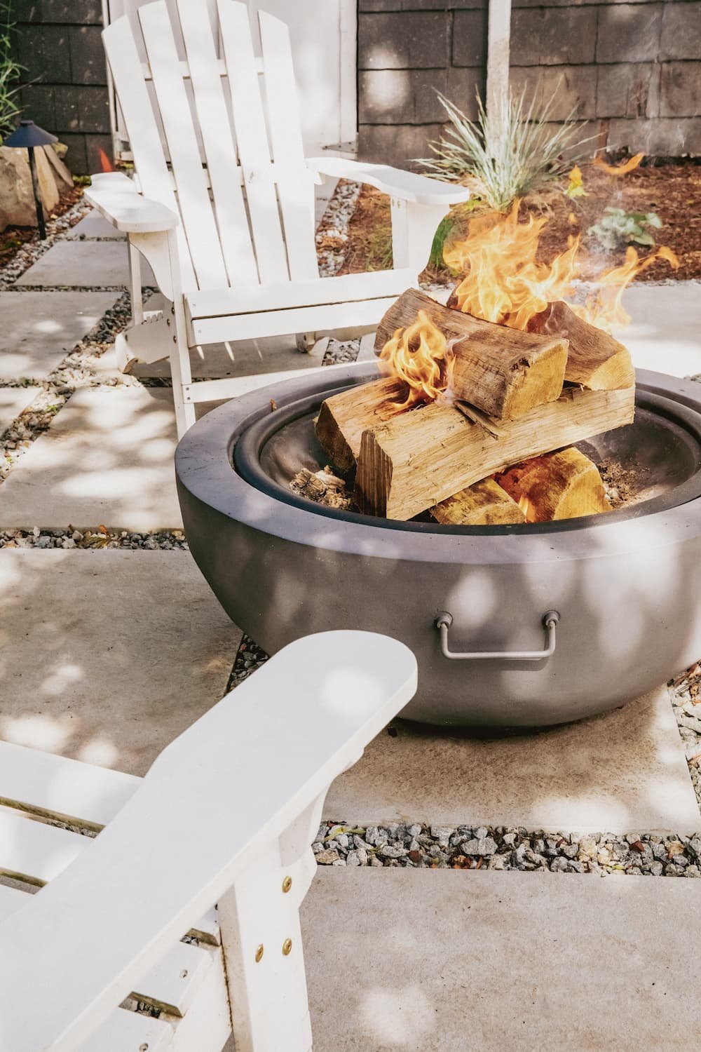 Close up of wood fire burning in modern bowl-shaped concrete fire pit and white adirondack chairs on concrete paver patio