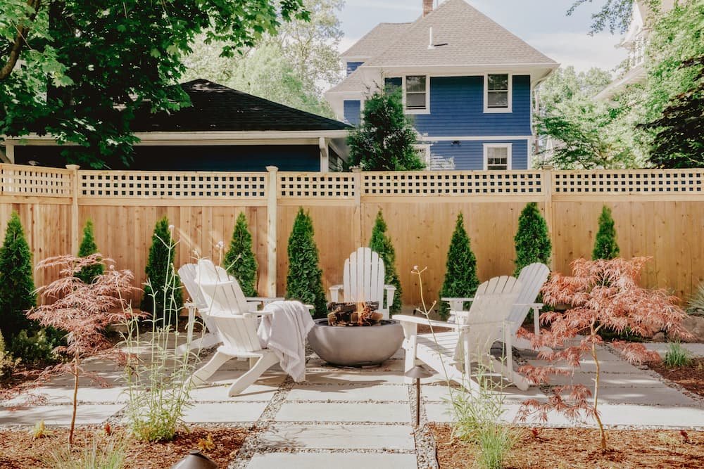 wide view of modern fire pit area on concrete paver and gravel patio with new surrounding fence flanked with tall evergreen planting