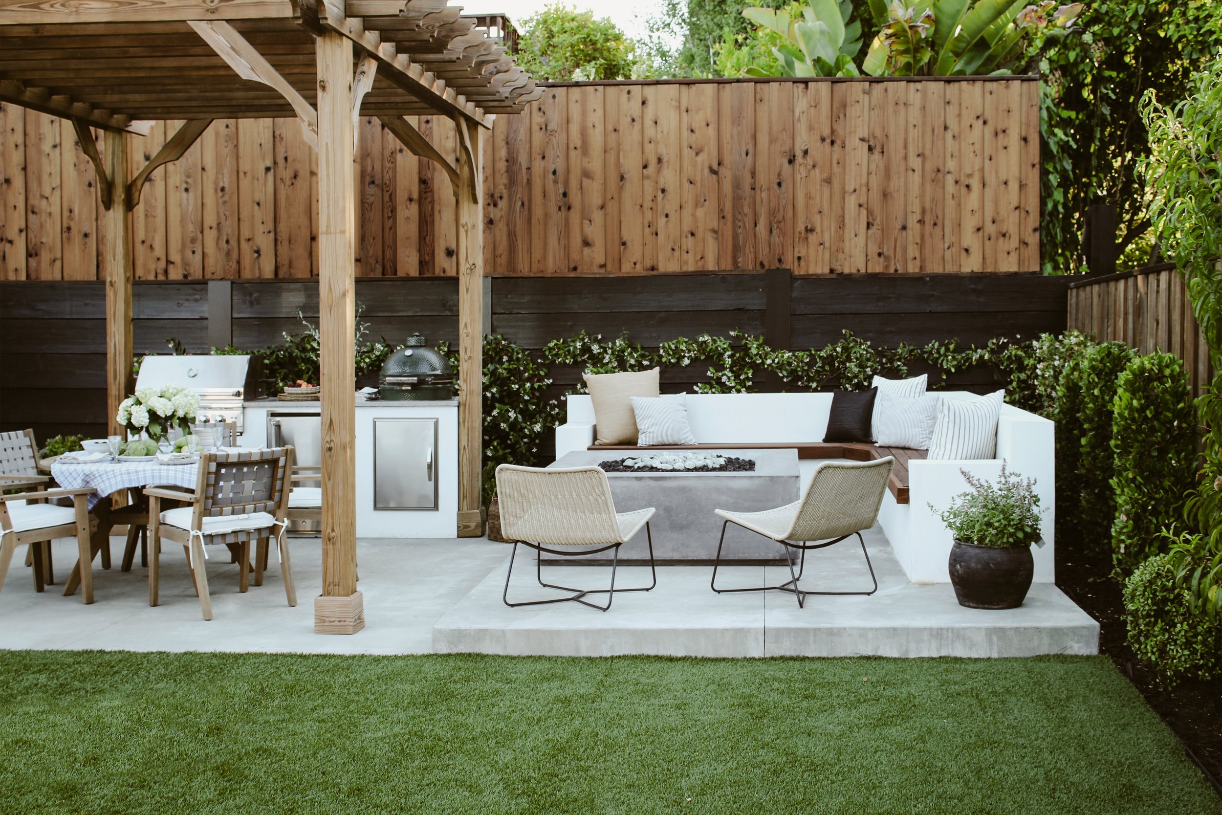 View of corner of finished backyard with L-shaped bench around a fire pit and pergola-covered dining table and outdoor kitchen