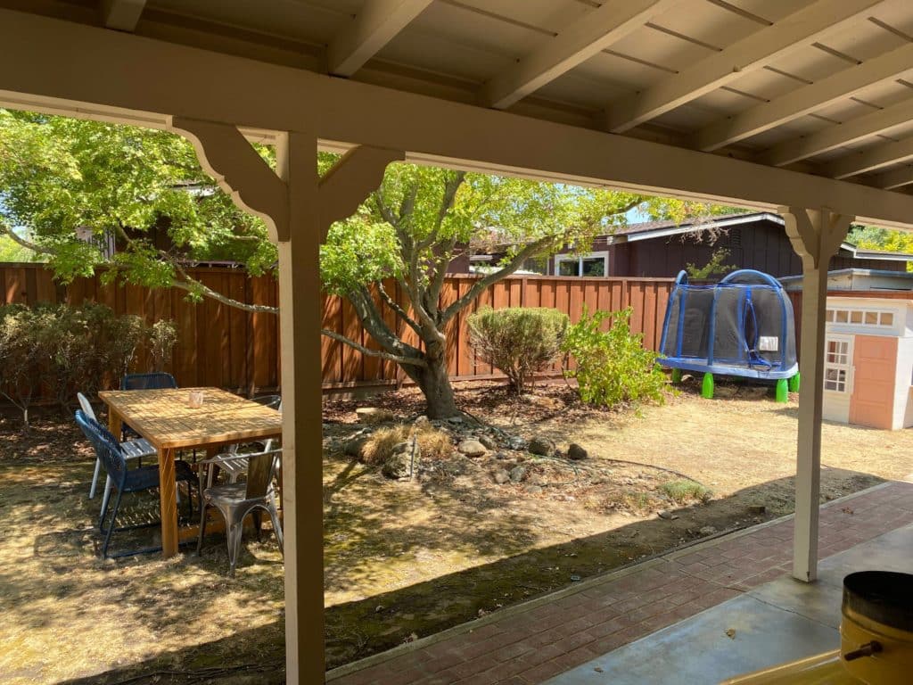 View of uninspired backyard from patio with thriving tree surrounded by dead lawn and haphazardly placed outdoor dining set and kids playset